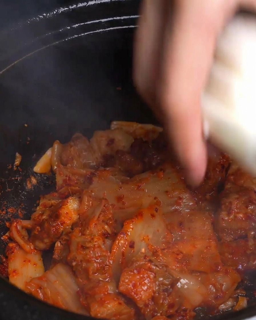 Una mano arrojando rodajas gruesas de cebolla blanca fresca a una olla caliente y humeante de kimchi rojo y panceta de cerdo salteados.