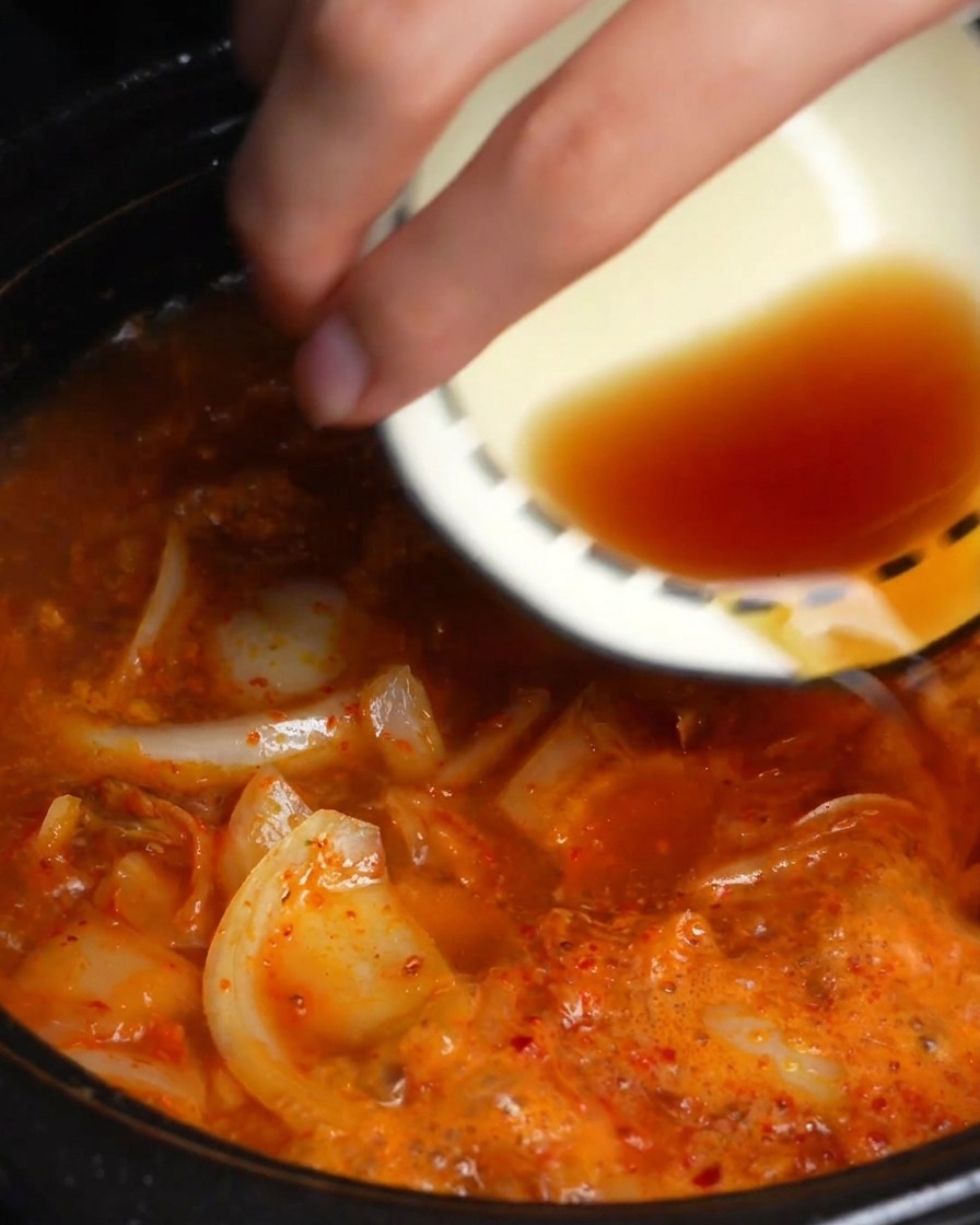 Una mano vertiendo soja oscura y salsa de pescado de un pequeño tazón en un caldo rojo de kimchi hirviendo a fuego lento.