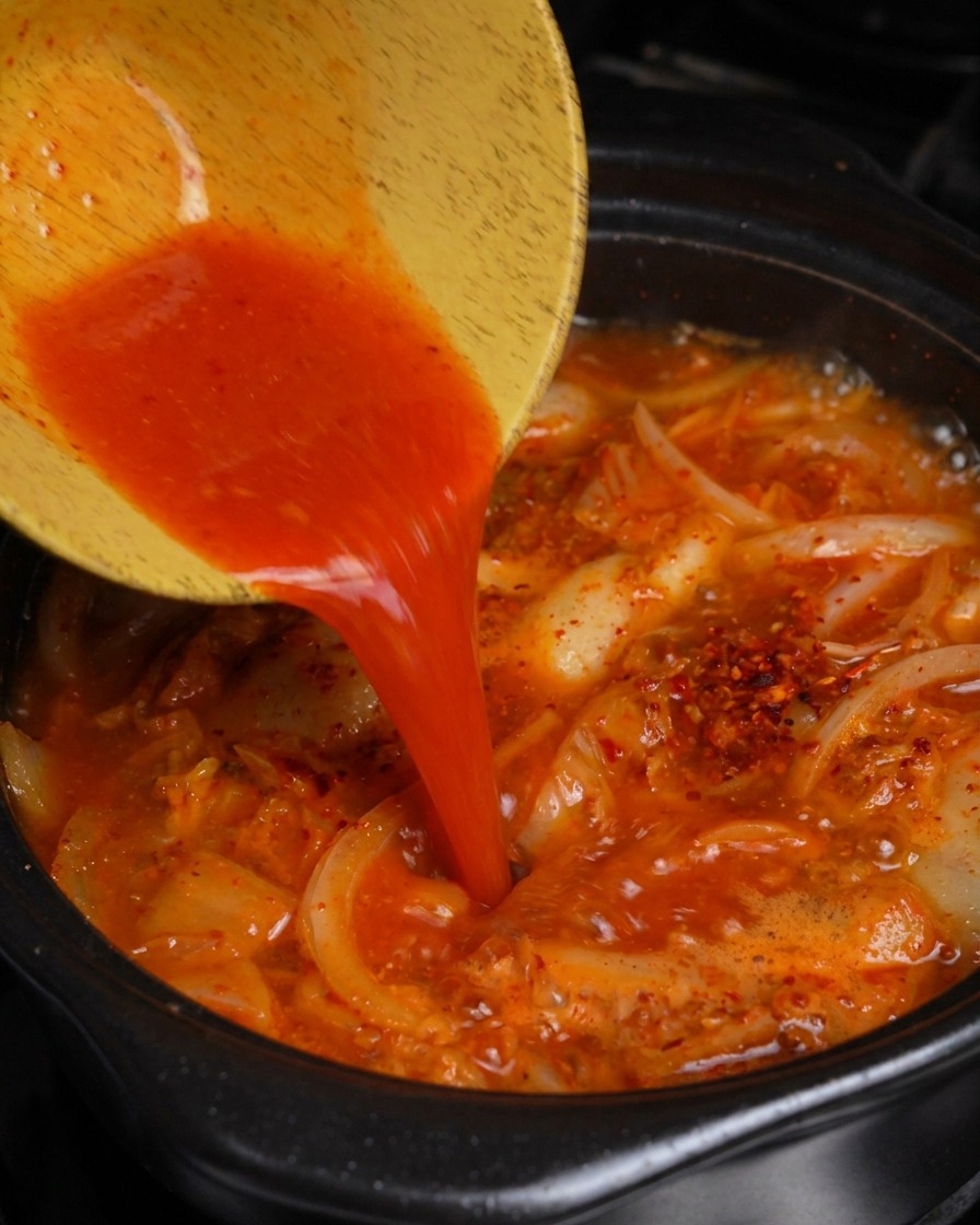 Vertiendo jugo de kimchi rojo brillante de un tazón de madera en una olla de estofado burbujeante de cerdo y kimchi.