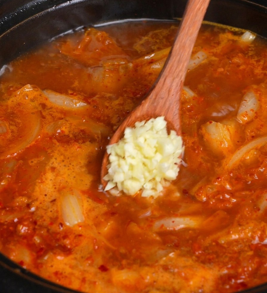 Una cuchara de madera agregando una porción de ajo fresco picado en una olla humeante de sopa de kimchi roja.