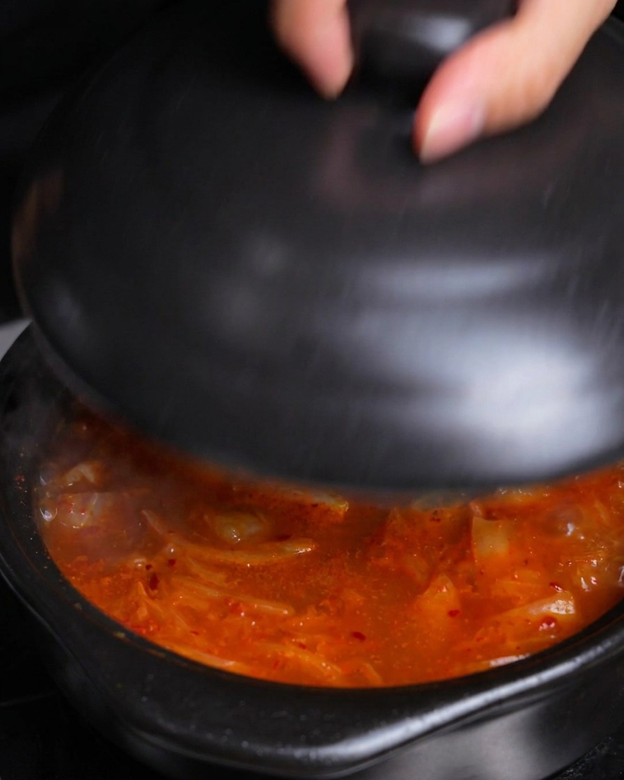 Una mano colocando una tapa de cerámica negra en una olla de estofado de kimchi burbujeante y caliente para dejarla hervir a fuego lento.