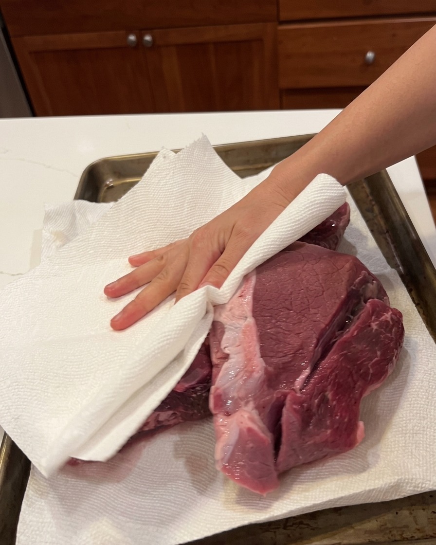 Una mano usando papel de cocina para secar un trozo grande de carne cruda en una bandeja.