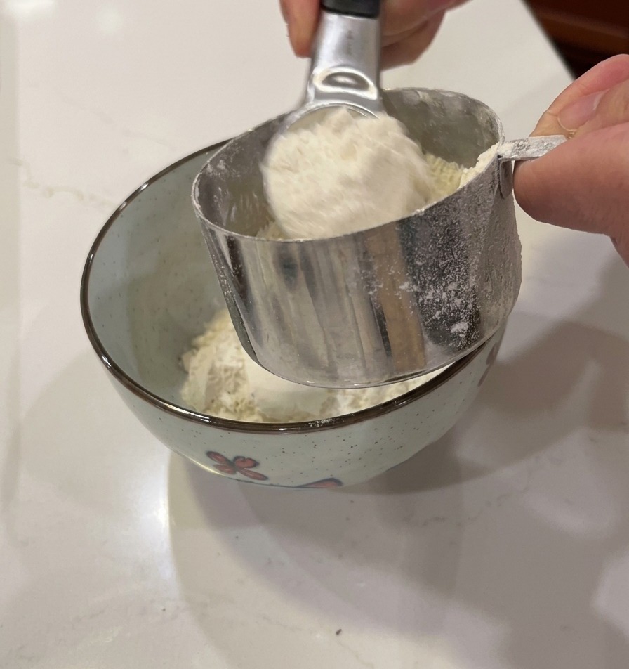 Manos vertiendo harina blanca desde una taza medidora en un pequeño bol decorado.