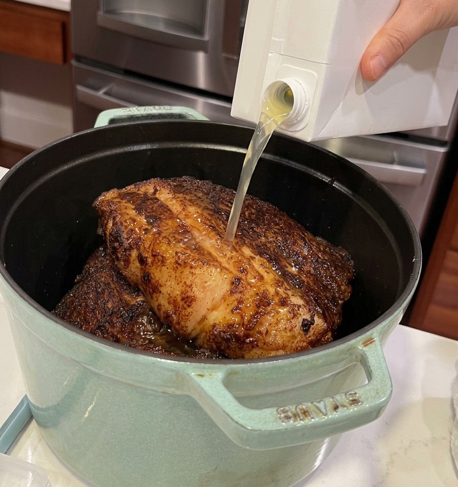 Vertiendo caldo de pollo desde un cartón en una olla de hierro verde llena de trozos de carne sellada.