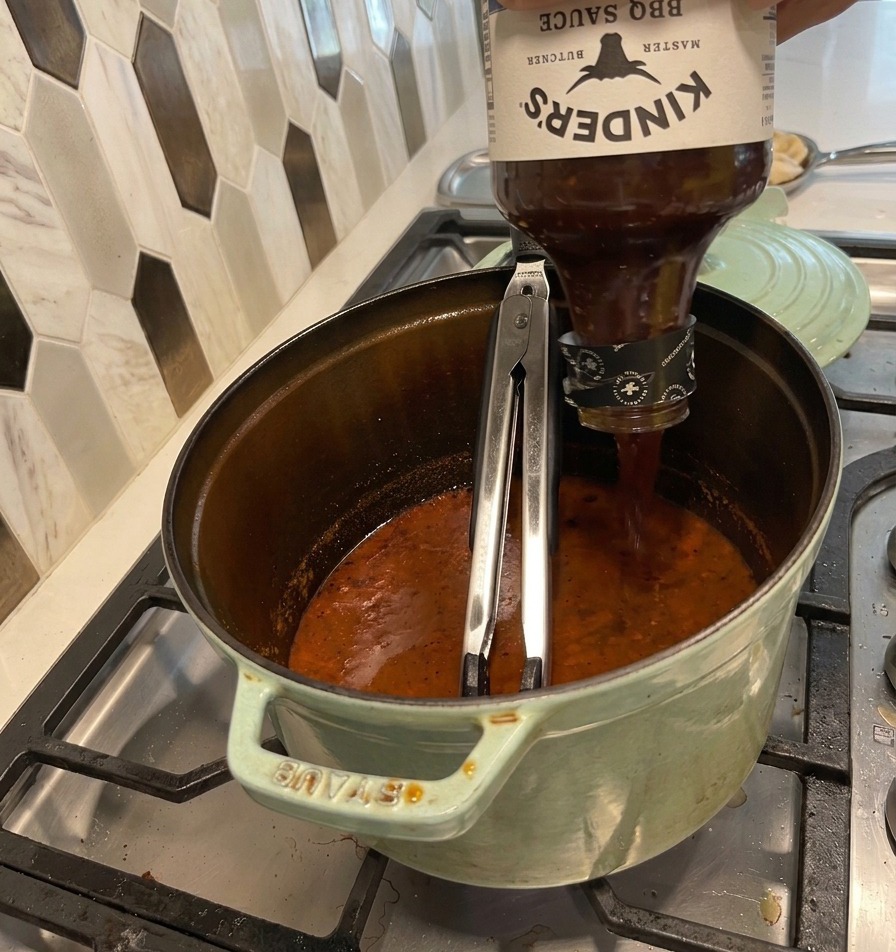 Vertiendo una botella grande de salsa BBQ en una olla verde con un líquido oscuro y reducido.