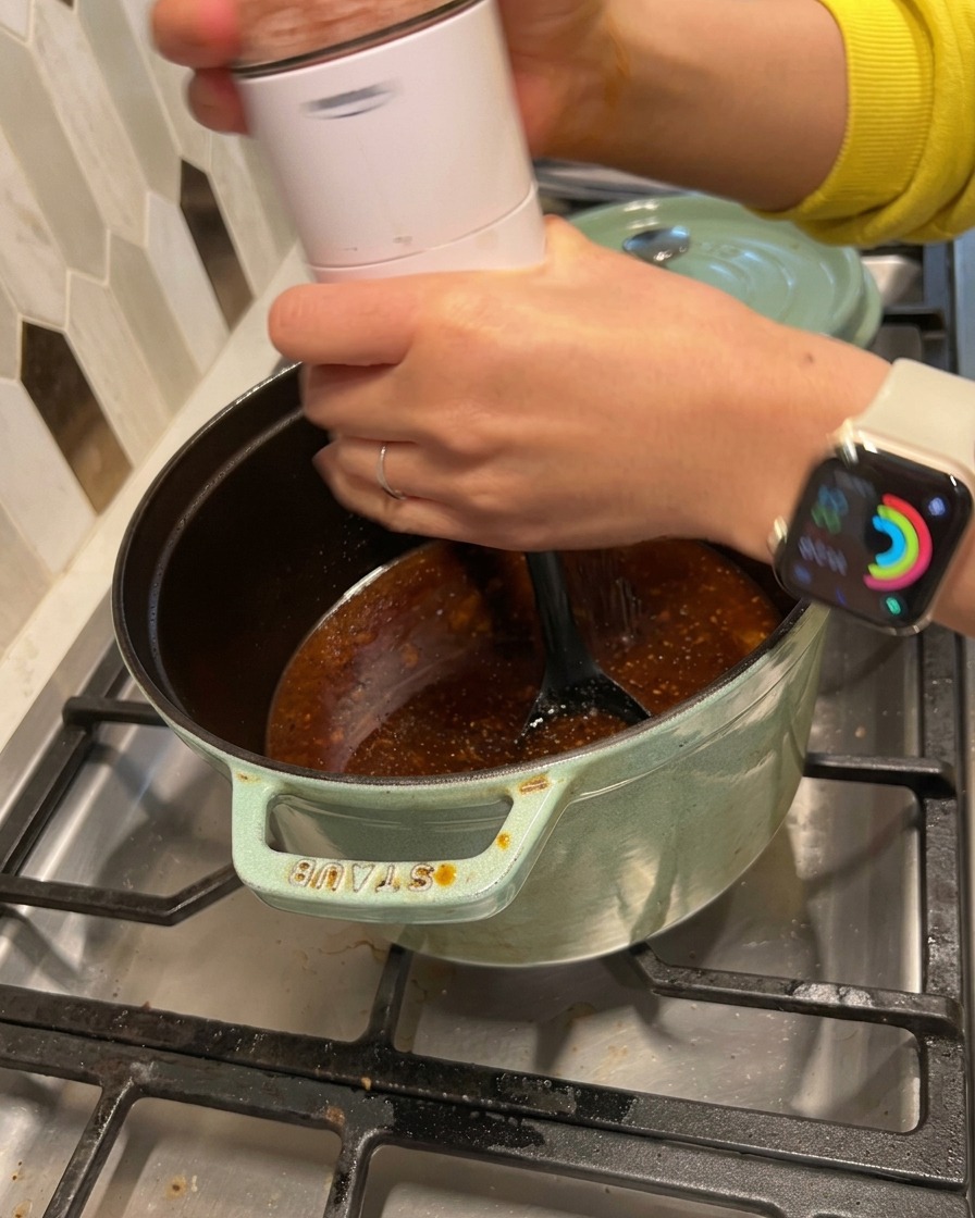 Una persona moliendo sal y pimienta negra en una salsa oscura que hierve a fuego lento en una olla de hierro.