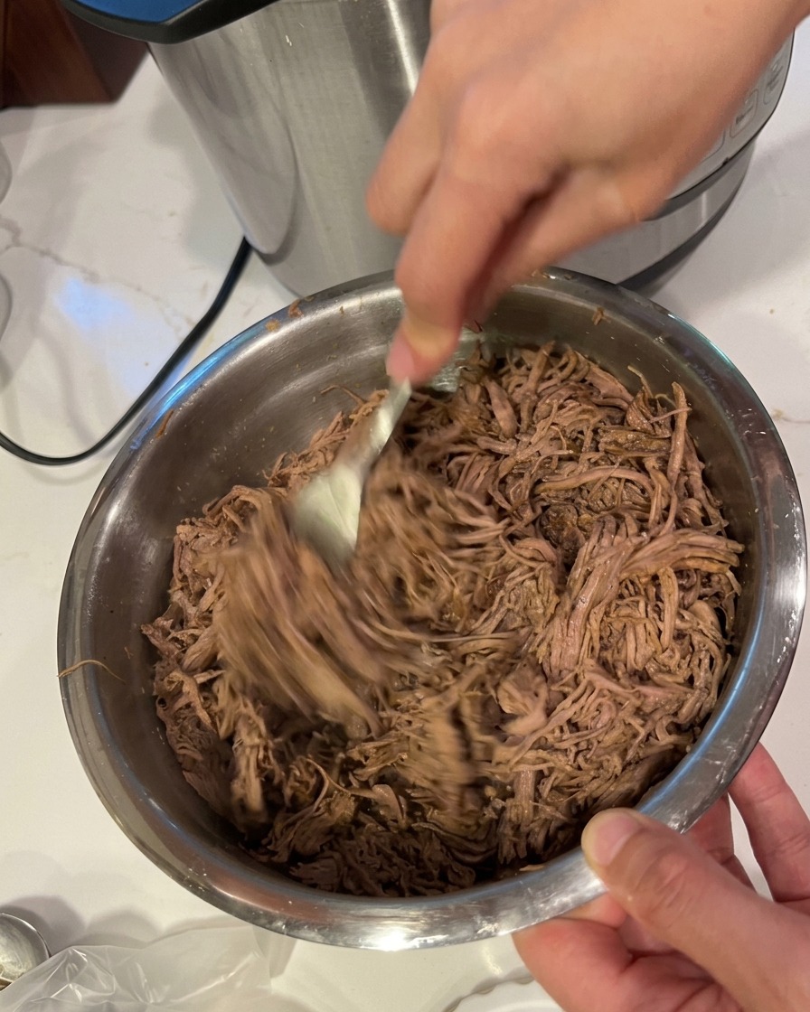 Dos manos usando tenedores para desmechar carne cocida en finas hebras dentro de un bol de acero inoxidable.
