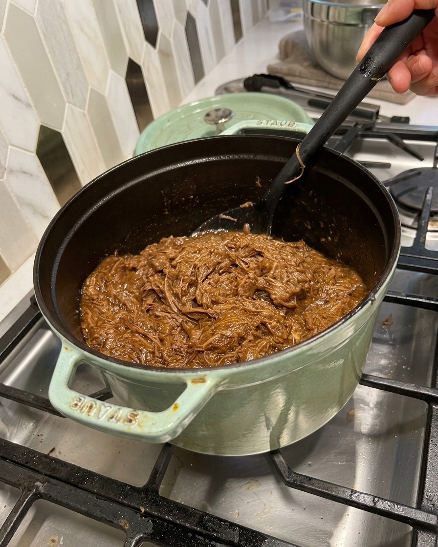 Carne desmechada siendo mezclada con una salsa barbacoa oscura en una olla de hierro con una espátula negra.