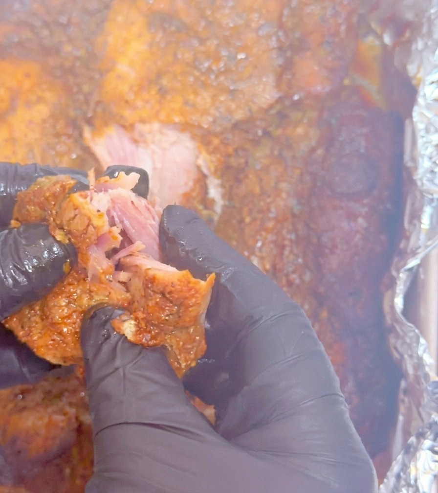 Manos con guantes negros desmenuzando un trozo tierno de cerdo cocinado a fuego lento, mostrando la textura suculenta.