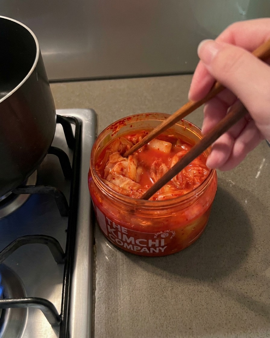 Palillos de madera sacando una porción de kimchi rojo y picante de un frasco transparente.