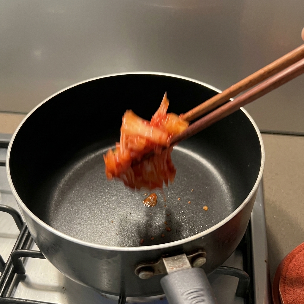 Kimchi rojo sazonado cayendo en una cacerola antiadherente oscura usando palillos de madera.