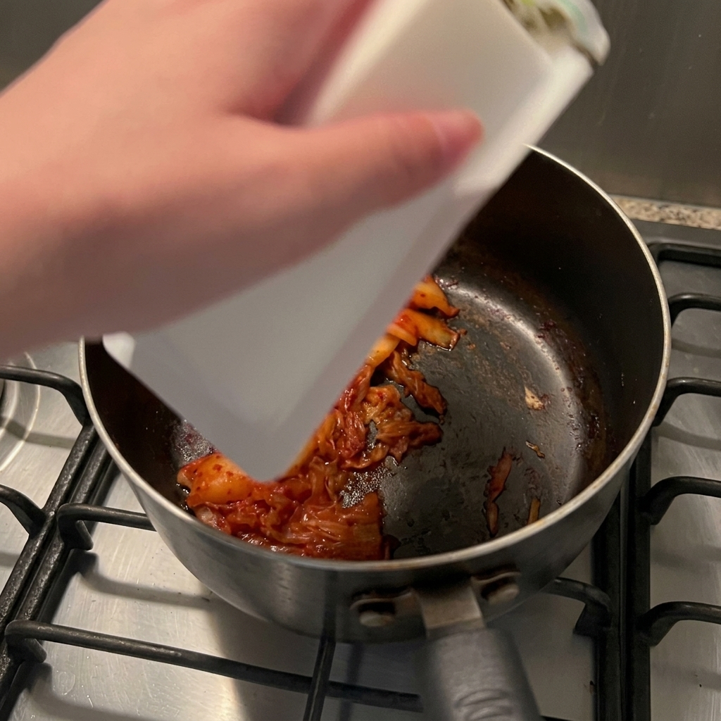 Un bloque de tofu suave blanco deslizándose desde su envase hacia una cacerola con kimchi salteado.