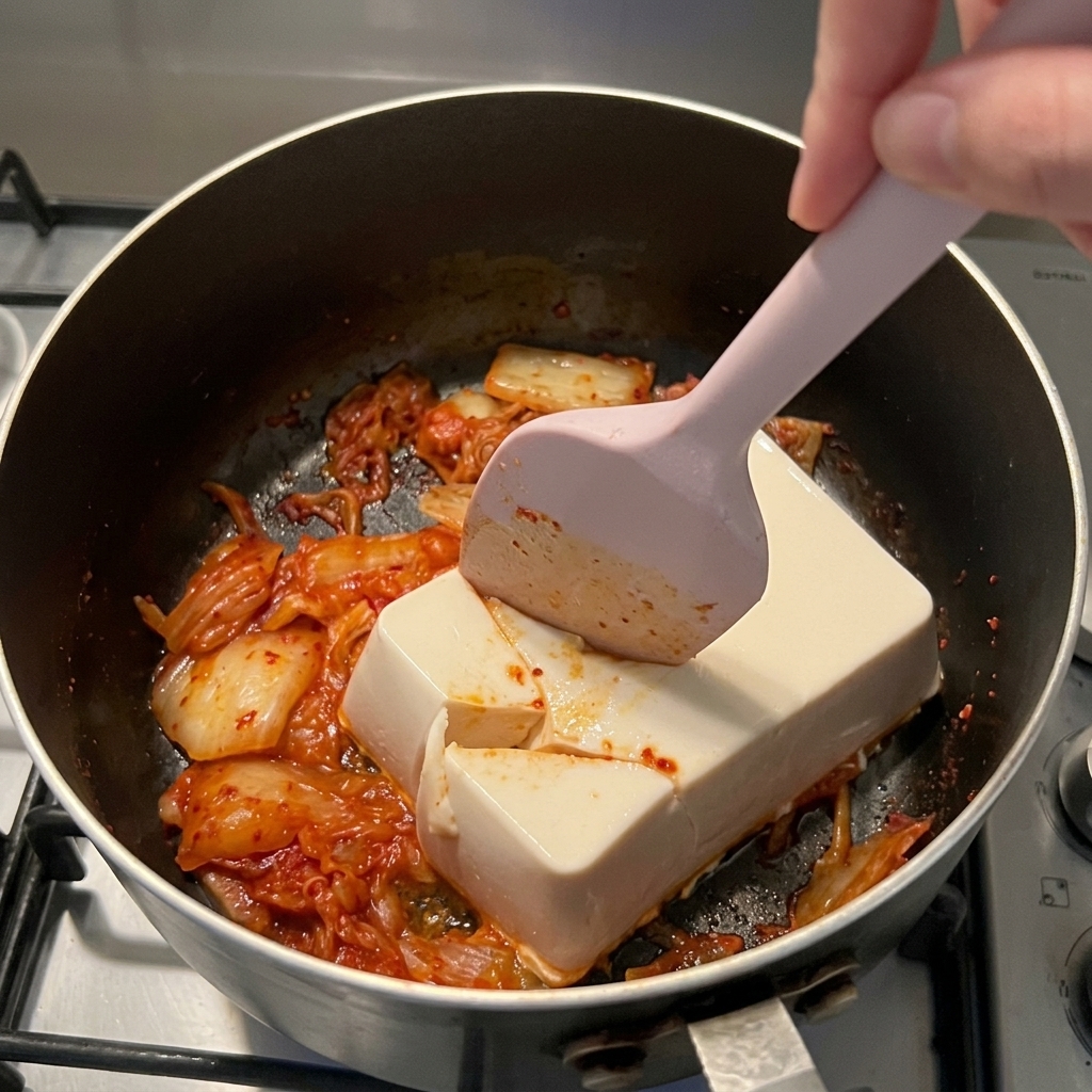 Una espátula de silicona rosa cortando un bloque de tofu suave sobre kimchi rojo en una cacerola.