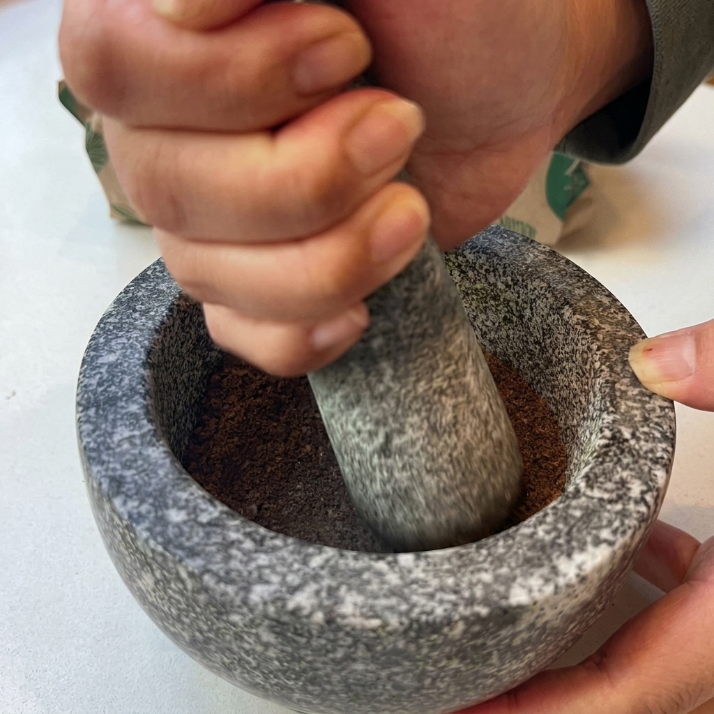 Manos usando un mazo de piedra para moler especias tostadas hasta obtener un polvo fino dentro de un mortero de granito.