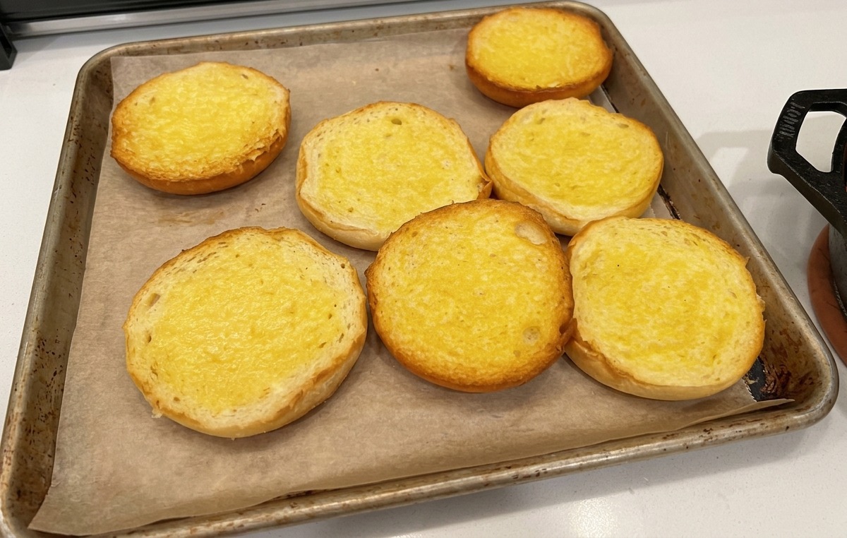 Mitades de panes de hamburguesa untadas con mantequilla sobre papel de horno en una bandeja de hornear.