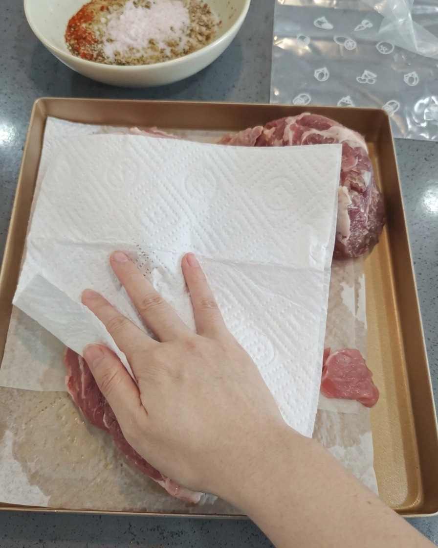Una mano usando papel de cocina blanco para secar trozos de cerdo crudo en una bandeja dorada.