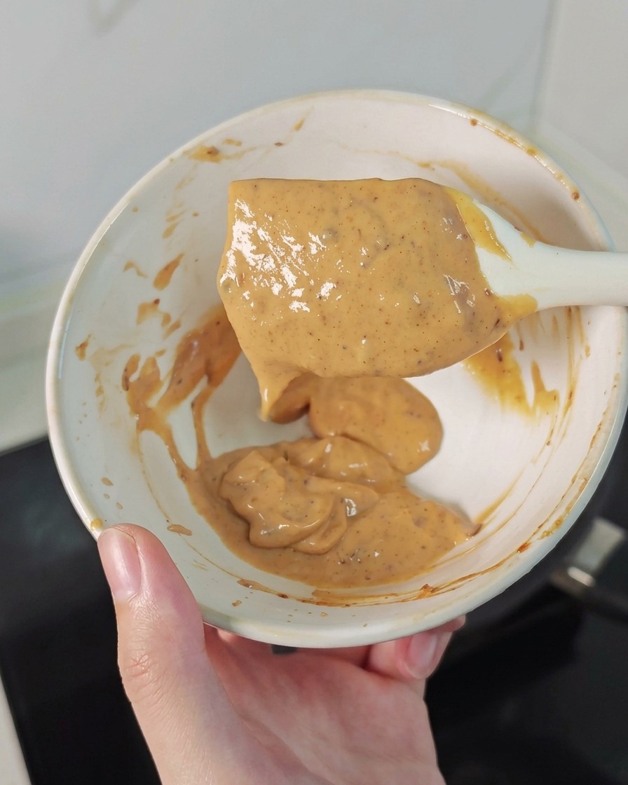 Una mano sosteniendo un bol pequeño y una espátula con una salsa cremosa de color naranja moteado.