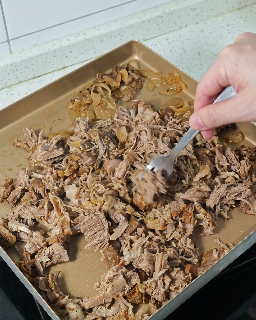 Una mano usando un tenedor para desmenuzar trozos de cerdo cocinado en una bandeja dorada.