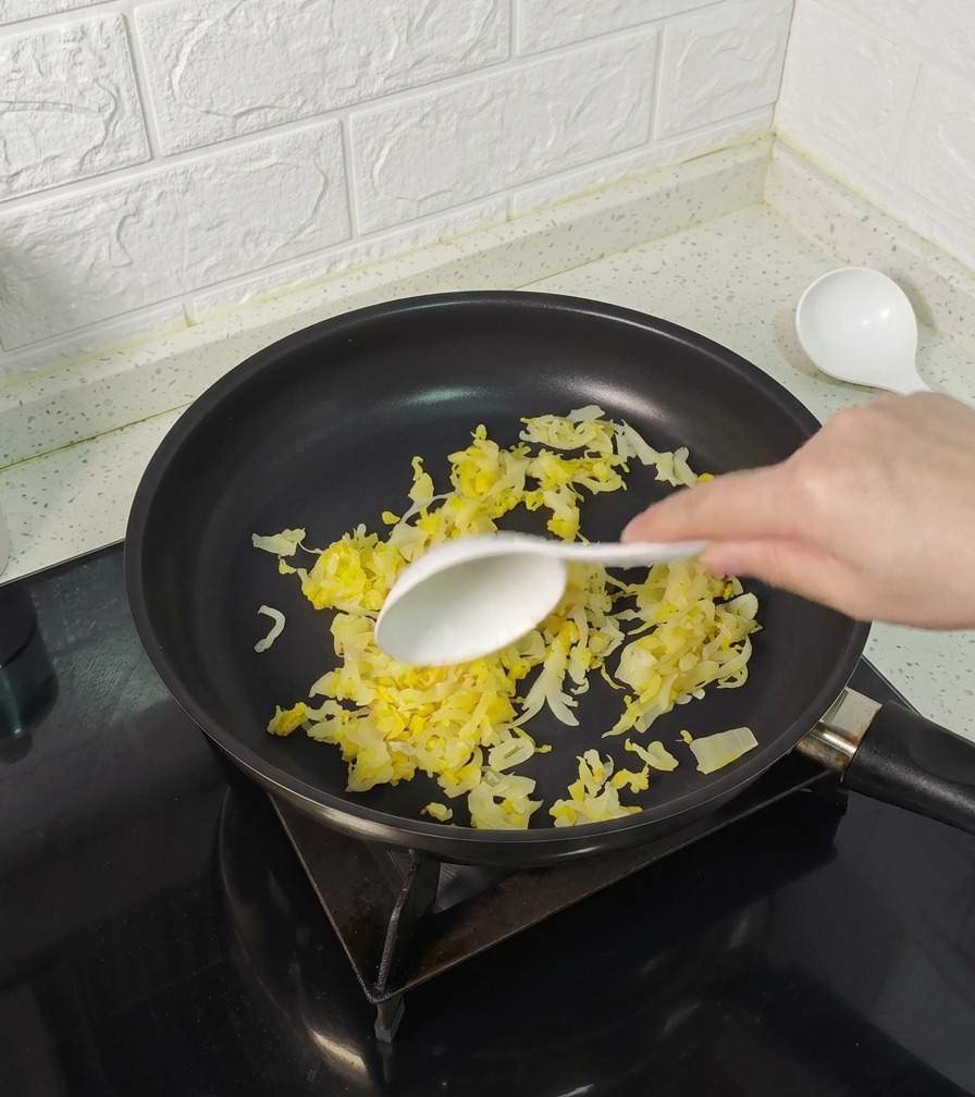 Una mano usando una cuchara blanca para remover chucrut amarillo en una sartén antiadherente negra.