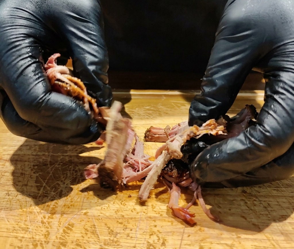 Manos con guantes desmenuzando una pieza de cabecero de lomo de cerdo asado a fuego lento sobre una tabla de madera.
