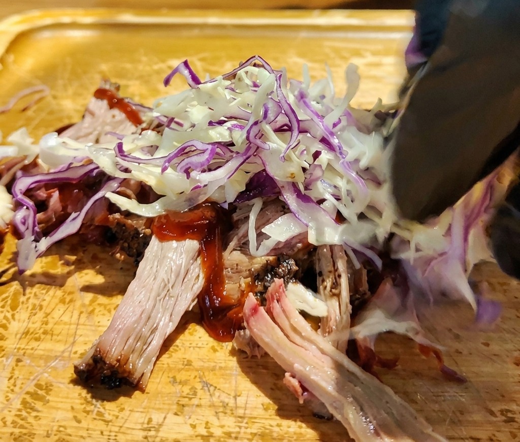 Mano colocando col roja y blanca rallada sobre el pulled pork con salsa en una tabla de cortar de madera.