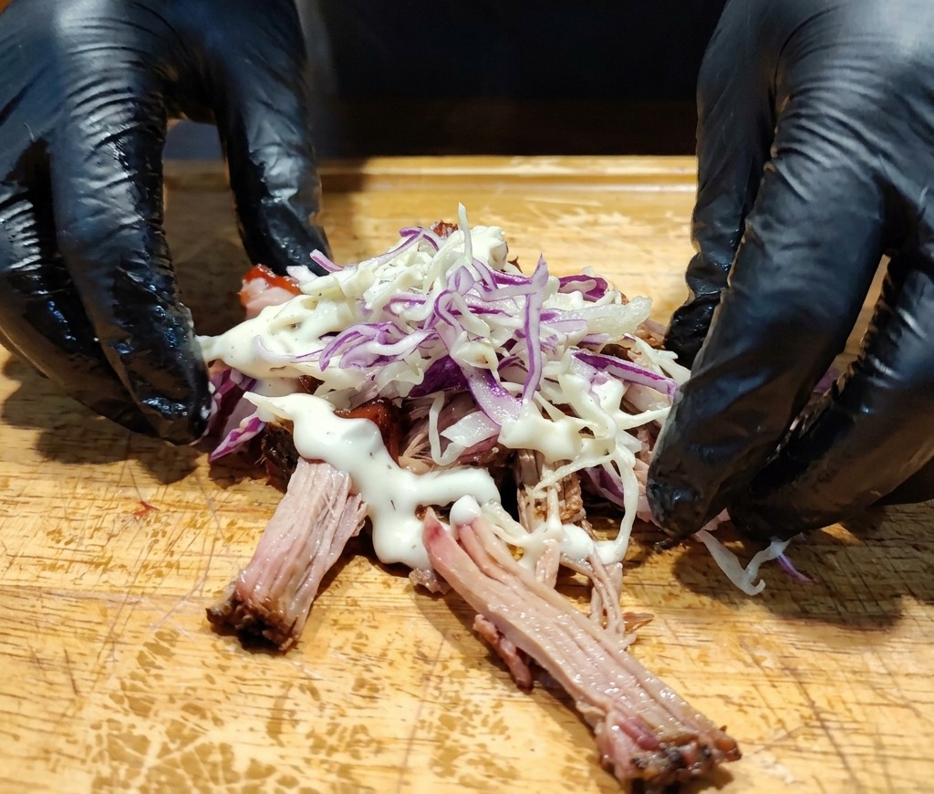 Manos junto a un montón de pulled pork cubierto con ensalada de col y un aderezo blanco cremoso sobre la tabla.
