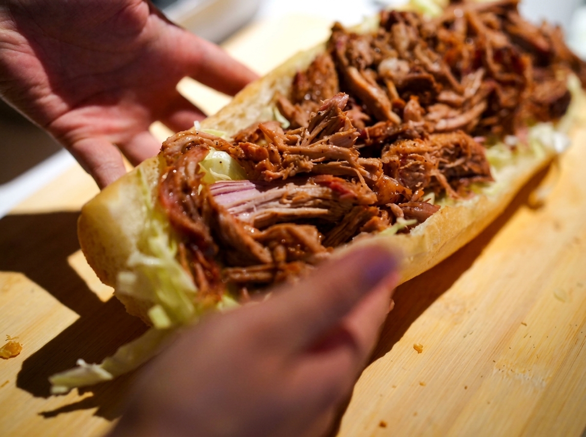 Manos sosteniendo un sándwich alargado relleno de ensalada de col y pulled pork con salsa.