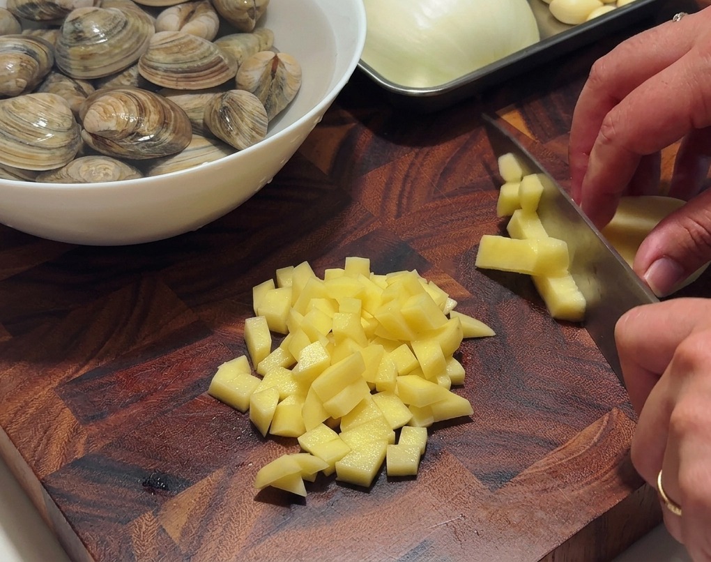 Manos utilizando un cuchillo de chef para cortar papas peladas en cubos pequeños sobre una tabla de madera.