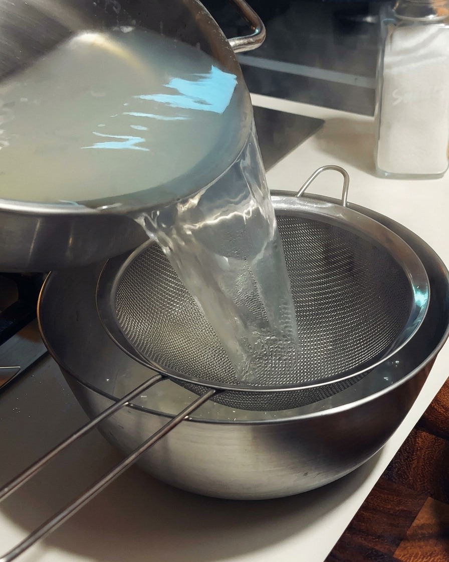 Caldo de almejas caliente y turbio siendo vertido a través de un colador de malla fina en un bol de metal.
