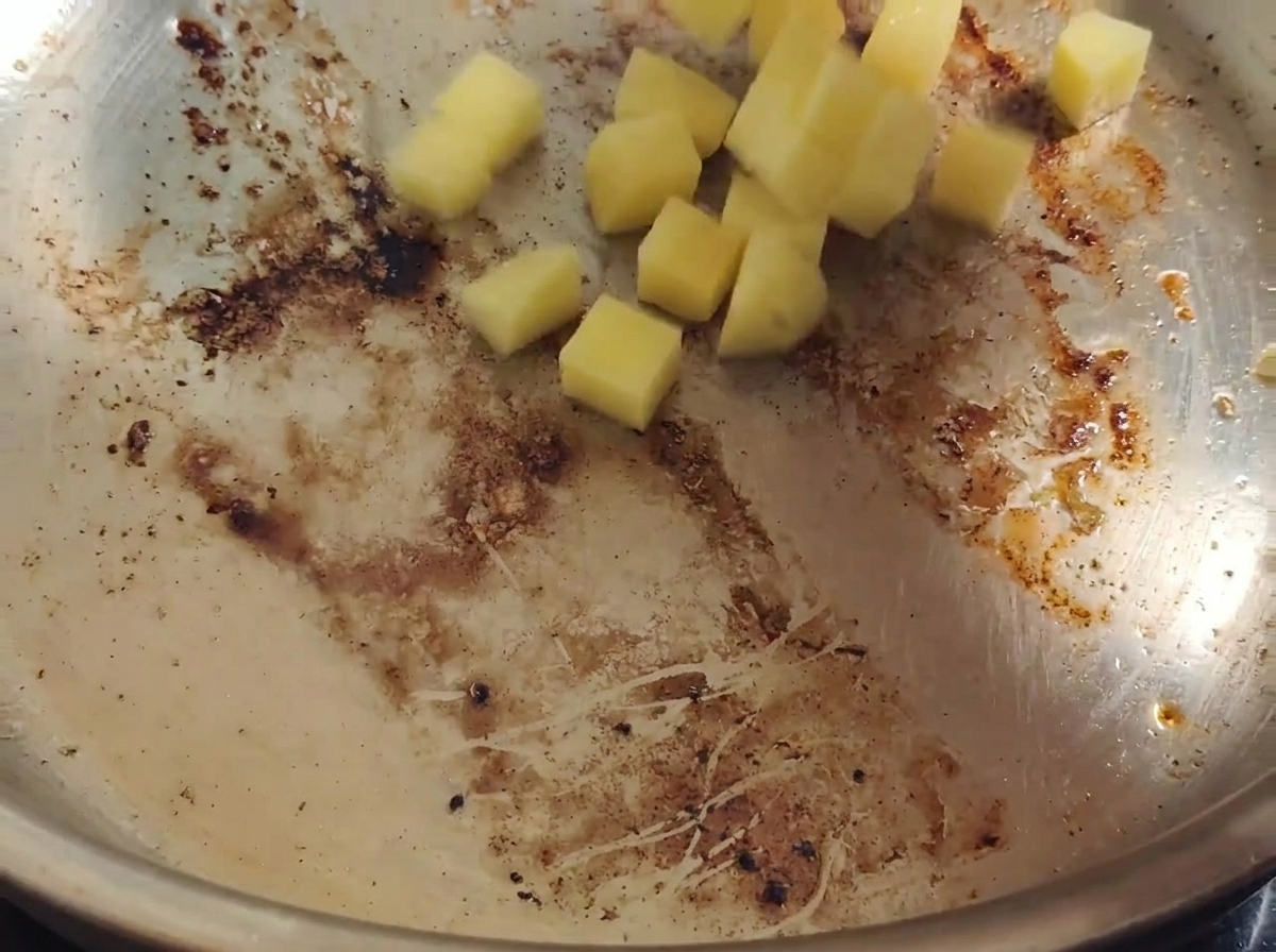 Papas amarillas en cubos cayendo en una sartén caliente con grasa de tocino y restos dorados.