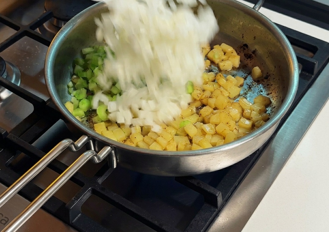 Cebolla blanca picada siendo añadida a una sartén con apio y papas doradas.