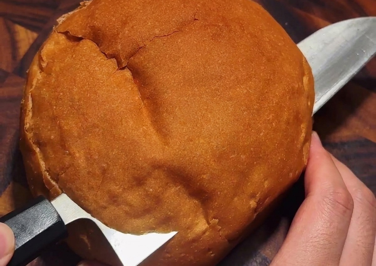 Un cuchillo cortando una tapa circular en la parte superior de una hogaza de pan rústico redonda.