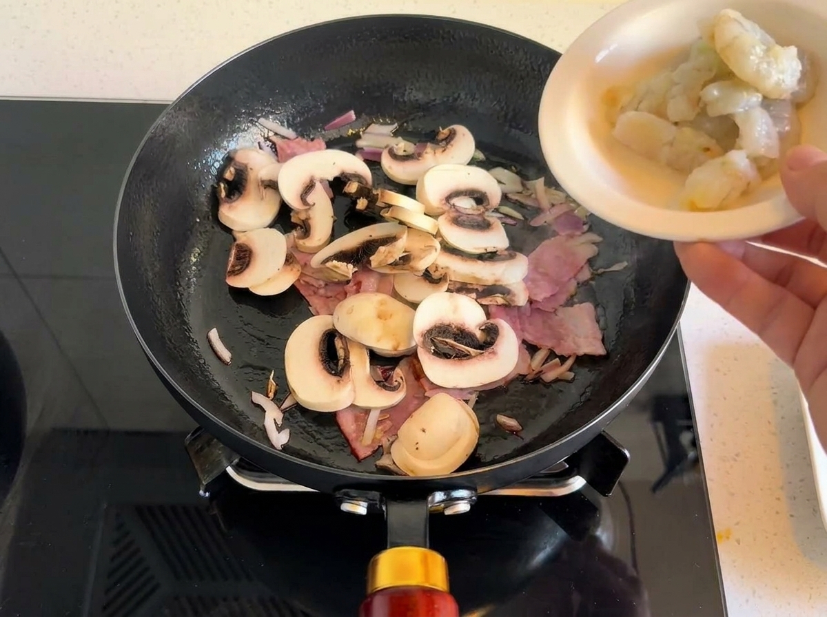 Camarones crudos siendo añadidos a la sartén con el salteado de tocino y champiñones.