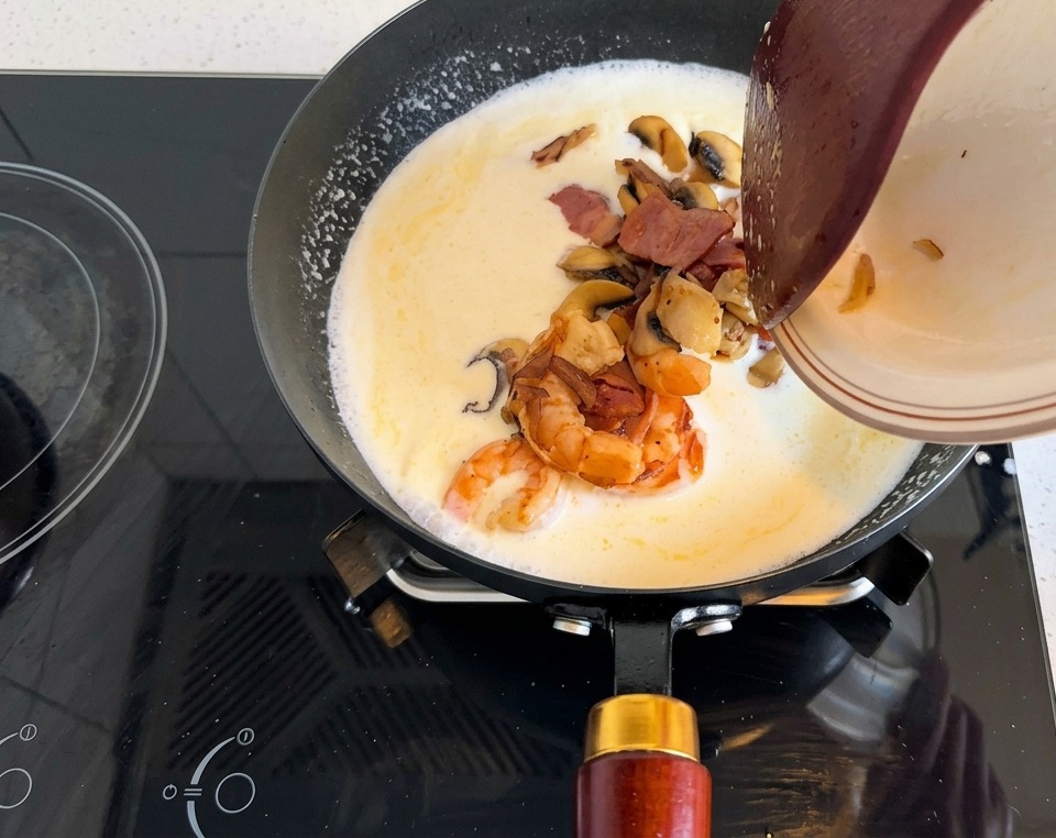 Plato con camarones, tocino y champiñones siendo vertido en la base cremosa de la sartén.