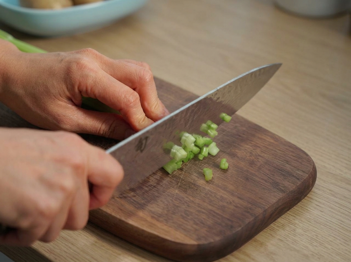 Manos usando un cuchillo de chef para picar ramas de apio verde brillante sobre una tabla de madera.