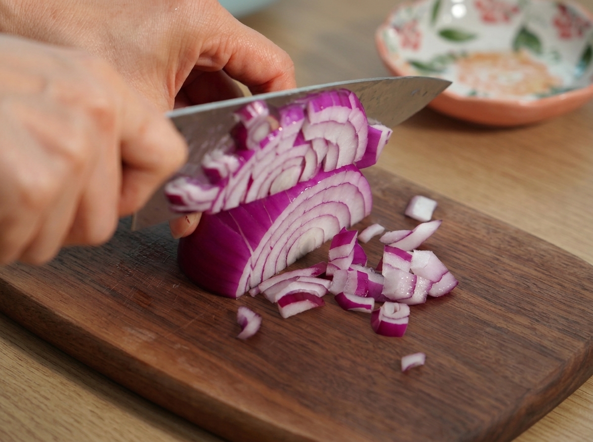 Manos usando un cuchillo de chef para picar una cebolla roja en trozos finos sobre una tabla de madera.