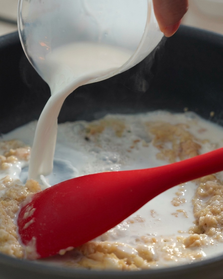 Leche vertiéndose desde una jarra medidora en una sartén con una pasta espesa de mantequilla y harina, removida con una espátula roja.