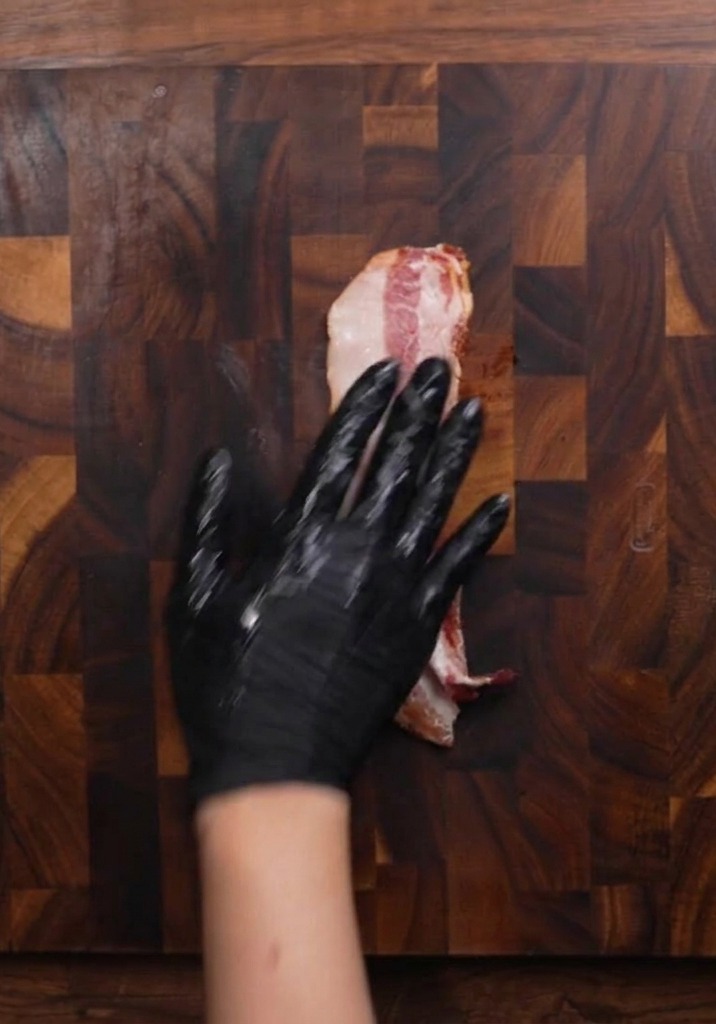 Mano con guante cortando tiras de bacon crudo sobre una tabla de madera oscura.