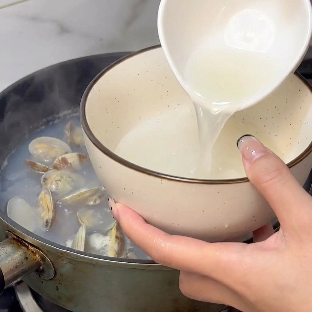 Una mano usando un cuenco blanco para recoger el caldo humeante de las almejas de la sartén.