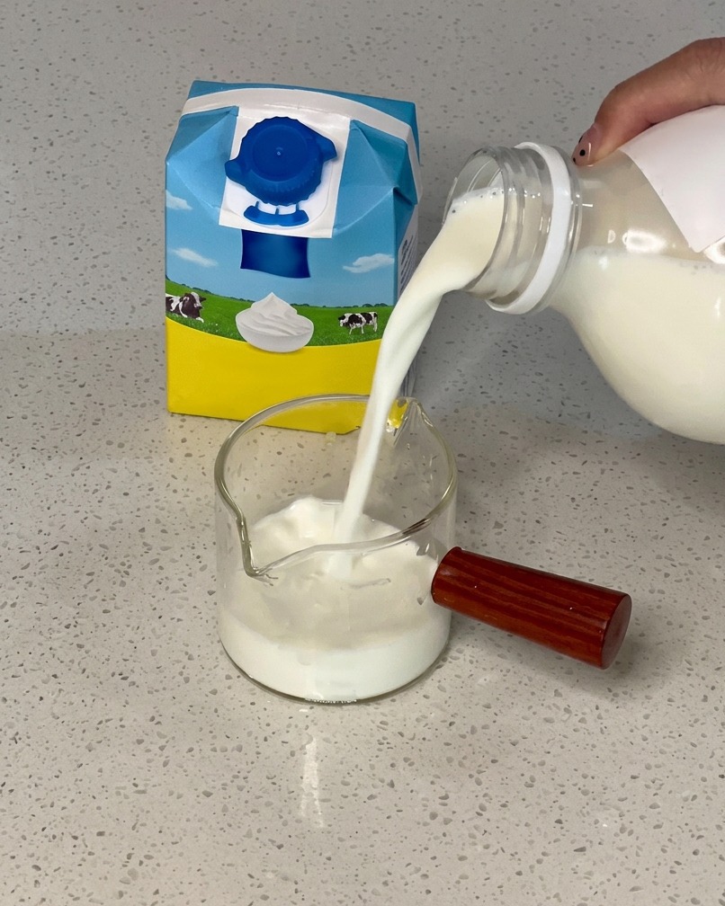 Mano vertiendo leche desde un brick azul y amarillo en una jarra de cristal con nata.