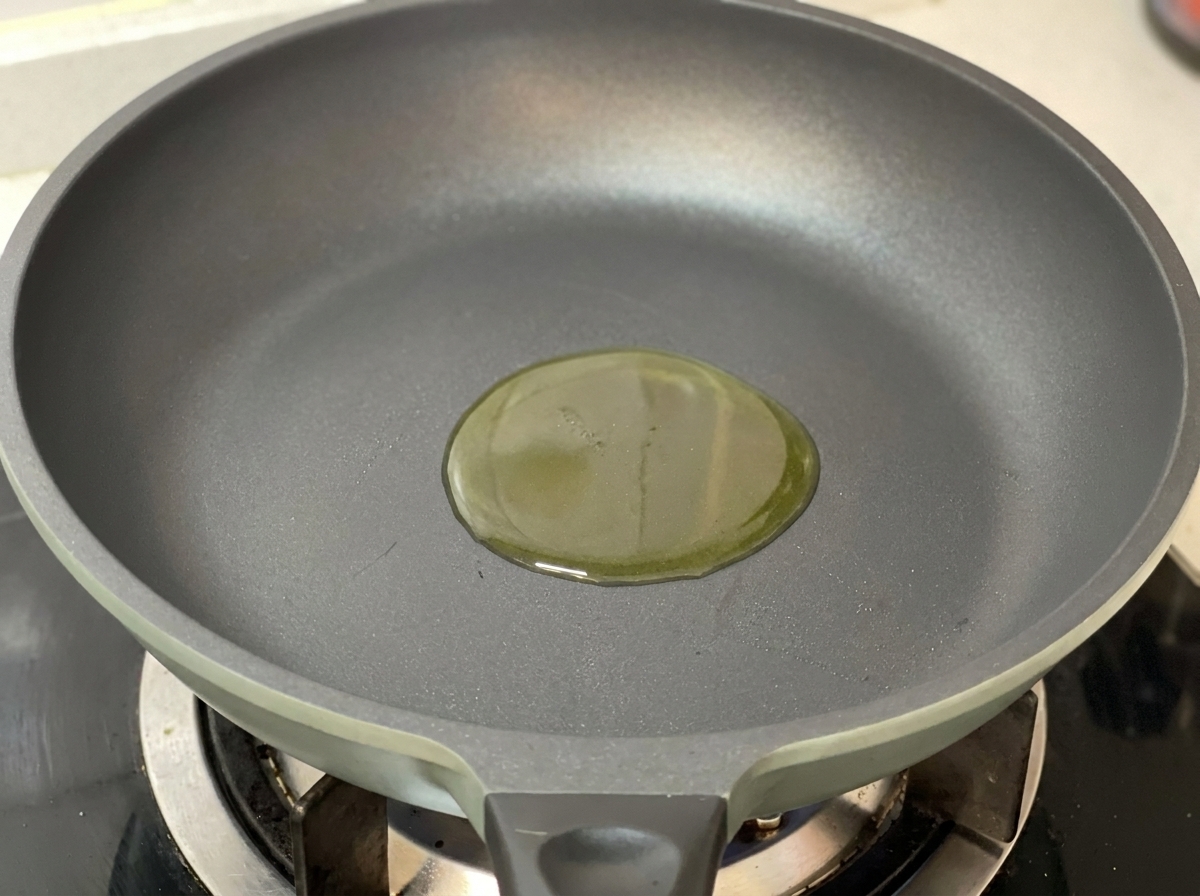Un pequeño charco de aceite de oliva dorado en el centro de una sartén gris sobre un fuego de gas.