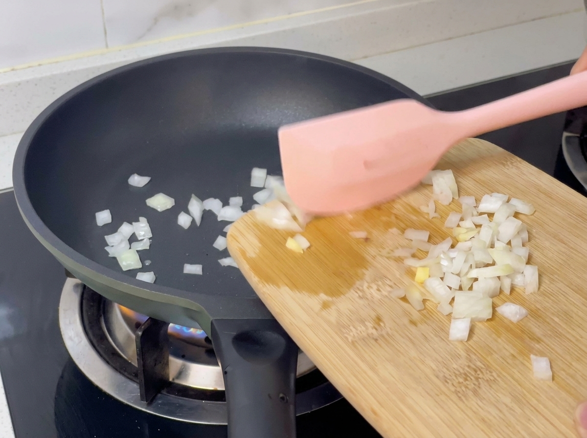 Cebolla picada cayendo desde una tabla de madera a una sartén caliente con la ayuda de una espátula rosa.