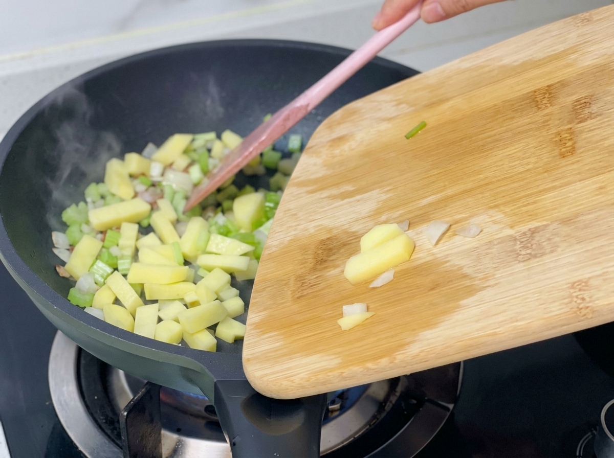 Dados de patata añadiéndose a la sartén que ya contiene apio, cebolla y bacon.