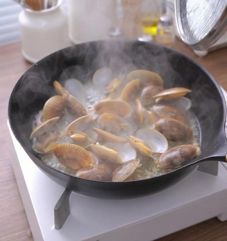 Vapor saliendo intensamente de una sartén con almejas cocinadas con las conchas abiertas.