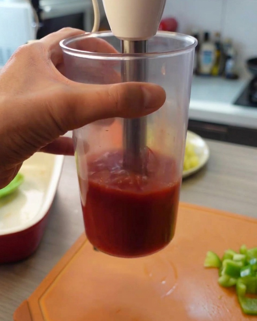 Usando una batidora de mano para hacer puré los tomates enlatados dentro de un vaso alto de plástico transparente.