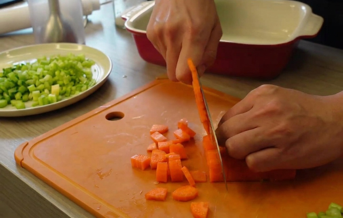 Manos usando un cuchillo de cocina para cortar zanahorias naranjas en pequeños cubos sobre una tabla de cortar naranja, con un plato de apio picado de fondo.