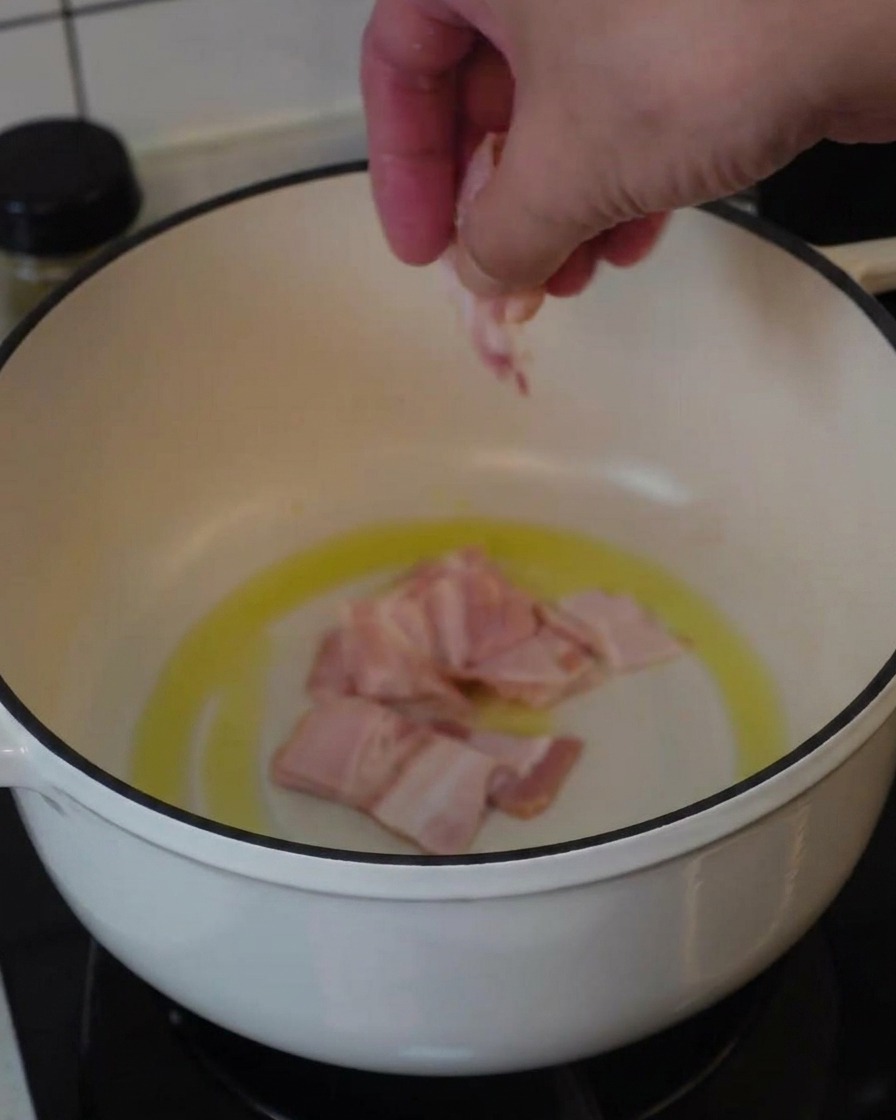 Una mano colocando trozos de tocino crudo en una olla blanca con aceite para empezar a freír.