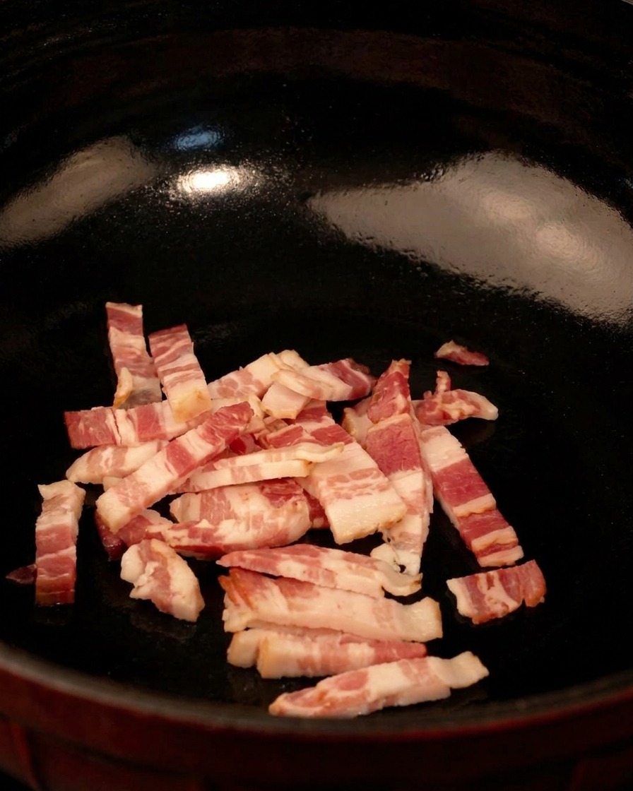 Tiras de tocino crudo en cubos en una sartén negra, listas para cocinarse a fuego medio.