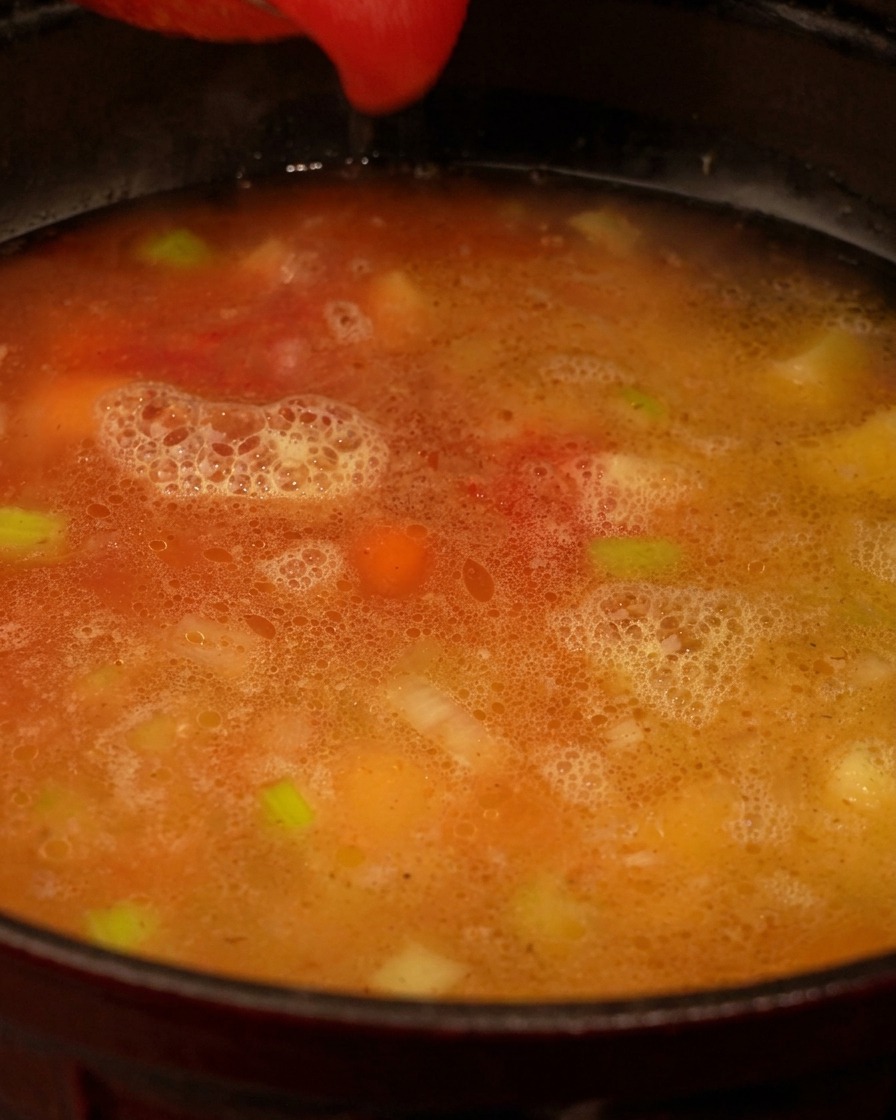 Puré de tomate rojo siendo vertido en una olla que contiene un caldo claro con trozos visibles de apio y zanahorias.