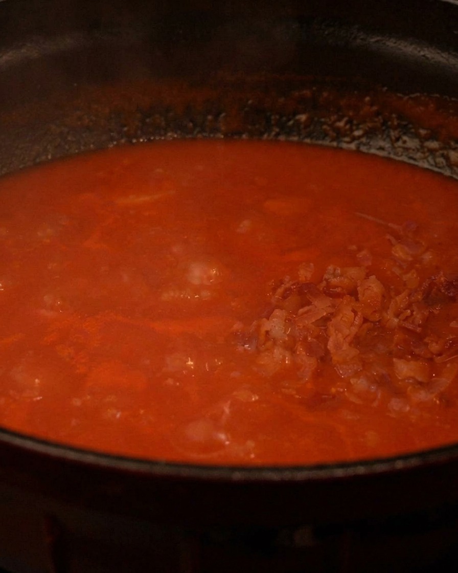 Trocitos de tocino crujiente siendo añadidos a una olla burbujeante de rica sopa de tomate roja.